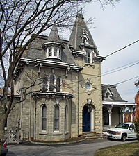 An ornate grayish-brown house with peaked and pointed towers with a bare tree at the left and a white car in a driveway on the right in front of it.