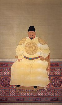 A middle aged bearded man wears yellow robes with dragons inscribed and a black hat, and sits on a throne.
