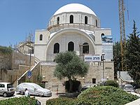A building covered in shimmering white Jerusalem stone stands against a backdrop of a clear blue sky. A bright white dome roof, supported by a bricked parapet including several arched unglazed windows, sits above a stone arch. The stone arch spans the width of the building and three arched unglazed windows lie below it. They are flanked by two towers on either side. Scaffolding is fixed around various parts of the external structure, with a scaffold staircase to the left of the building and the tall latticed boom of a tower crane on the right. The construction company’s promotional hoarding can be seen affixed to the right side of the building.