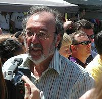A video camera is being pointed at a bearded man who is wearing glasses. Some other people stand in the background.