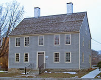 A light blue wooden house with a pointed gray roof, two white chimneys and pale cream-colored window and door trim