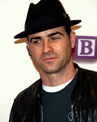 A black-haired man wearing a black fedora, a black leather jacket and a green T-shirt.