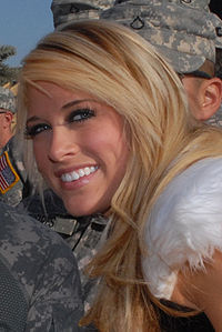 A close-up photo of a young, Caucasian blonde, who is smiling at the camera. Men in camouflage uniforms are visible in the background.