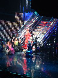 A woman wearing a red ensemble, standing in front of a metallic staircase around a group of dancers.