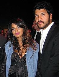 A man and a woman standing together. Both black hair and dark skin. The woman is wearing a light blue jacket over a black dress, and the man is wearing a white shirt and a black jacket.