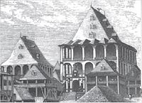a cluster of rectangular wooden buildings with peaked rooftops, the largest of which crowns the hill and is surrounded by wooden verandas on each of its three stories