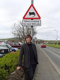 Mark Steel standing under a road warning sign in Kirkwall, Orkney, Scotland, which is warning people of "Otters crossing for next 600 yards."