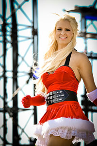 A Caucasian woman with blonde hair smiles while wearing a red dress with a white fur trim, in the style of Santa Claus. She is wearing elbow-length red gloves, also with a fur trim, and a black belt around her waist.