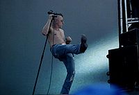 A man with a microphone stand holds his right leg into the air while facing to his left.