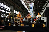Four males standing at microphones with three playing guitars, second male from left at keyboards, all are inside an industrial shed with skylights behind.