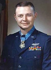 Head and shoulders of a man with short hair and a round face in a dark blue military jacket. Rows of ribbon bars and a winged pin are on his left breast and a medal hangs from a light blue ribbon around his neck.