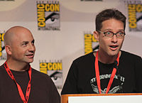 A man with a bald head and a brown sweater, and a man with spiked brown hair and glasses, speaking into a microphone.