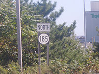 A road sign reading north above a black rectangle with a white oval containing 185 in black numberals. Trees and industrial buildings can be seen in the background.