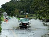 A pickup truck leaving high spray columns behind as it crosses a torrent of water crossing a two-lane road. Behind it is a line of vehicles waiting to make the same crossing