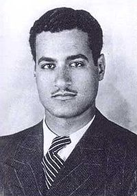 A man wearing a tweed jacket and a tie. His hair is raised and black and he has a mustache.