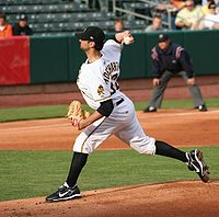 A side view of a young man in a baseball uniform, cap, and glove as he is about to throw a baseball off a pitcher's mound in a stadium. A uniformed umpire in the background looks on as the man has one foot nearly off the mound and the baseball behind his head.