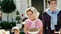  Image of a blond woman. She is a housewife and carries a pie in her left hand. Her hair is styled in 1950's fashion. She is wearing a pink polo shirt. Surrounding her are her husband and children.