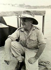 Informal portrait of man in light-coloured military uniform with wide-brimmed hat, crouching on one knee