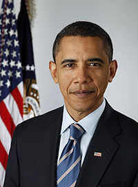 A portrait shot of a serious looking middle-aged African-American male looking straight ahead. He has short black hair, and is wearing a dark navy blazer with a blue striped tie over a light blue collared shirt. In the background are two flags hanging from separate flagpoles: an American flag, and one from the Executive Office of the President.