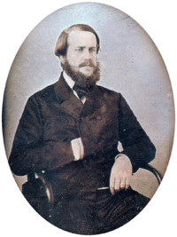 A photograph of a young bearded man seated in an armchair with his right hand tucked into the front of his double-breasted jacket