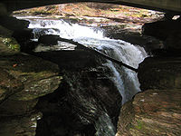 The upper half of a falls dropping into a narrow slot in layered rock, as seen from the side. The curved underside of a bridge is visible above the waterfall, which is in shadow.