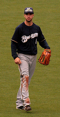Portrait of a man in a dark blue baseball jersey with "BREWERS" across the chest, grey pants with dirt on the right knee, a baseball glove on his left hand, and a blue cap with an "M" on it.