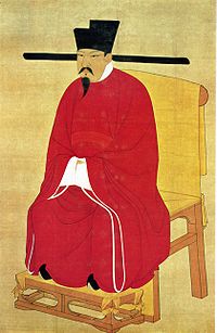 A portrait oriented scroll depicting a man with a pointy beard and mustache in thick red robes, black pointed shoes, and a square cut, black hat with long, thin protrusions coming out horizontally from the bottom of the hat, sitting on a throne.