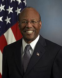 Undersecretary Stanley, wearing eyeglasses, dark blue suit with flag pin in his lapel