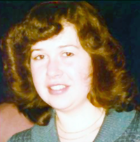 A slightly faded picture of a young woman with curly brown hair surrounding her slightly smiling face. She is dressed in a simple blue frock and has a simple necklace.