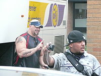 Two adult Latino males, one doing hand signs and another holding a camera.