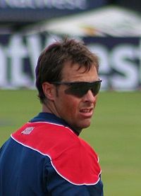 A man wearing sunglasses and navy blue and red English cricket t-shirt looks across over his shoulder at a sports ground. A sponsorship board is visible in the background.
