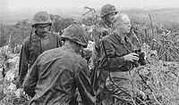 A black and white image of Merritt Edson and three other Marines standing in bushes. Edson is holding some binocculars and is looking out over the battlefield.