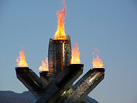 four pillars with flame at their tops surrounding a single fifth pillar in the middle, also with flame at the top. The background is sky with mountain.