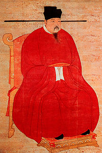 A portrait oriented scroll depicting a man in thick red robes, black pointed shoes, and a bucket shaped black hat with long, thin protrusions coming out horizontally from the bottom of the hat, sitting on a throne.