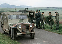 Group of three United States soldiers, one armed with a rifle, and two West German Bundesgrenzschutz officers standing by two vehicles parked on a narrow asphalted road in a rolling landscape with fields and hills visible behind them.
