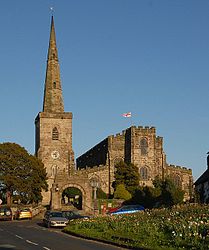 A stone church seen from the west showing, from the left. a tower and spire detached from the rest of the church, and the wide body of the church with a battlemented parapet