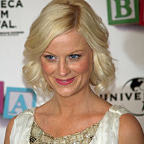 A shot from the shoulders up of a blond woman with blue eyes wearing a white and green dress, smiling and looking at something outside the image.