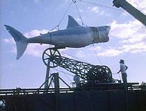 A large model shark is hoisted by a crane as two men watch it.