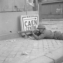 A soldier lies prone, rife at the ready by a building in a city street. Beside him is a sign reading "Caen centre", pointing back the way he has come.