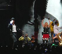A group of males and a female performing on stage in front of a crowd of people. A male is shown blowing a kiss to the audience while wearing a white glove on his right hand and a white shirt with black pants and a jacket. An African American male wearing a white and dark grey shirt is shown in a crouched down position. The female is waving her hands in the air while wearing black fingerless gloves with a black shirt and pink shorts. There is also two other African American males in the background making hand gestures. Behind the people on the stage, there is a screen that shows a black and white photo.