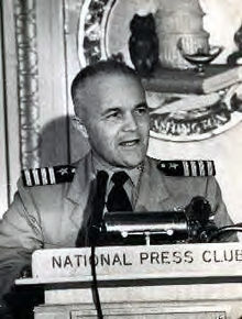 Middle-aged man wearing U.S. Navy khaki uniform and Captain shoulder epaulettes standing behind a lectern with microphone and placard that reads "National Press Club."