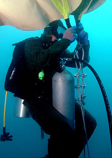 Underwater, a US Naval diver in a scuba suit w/mask, oxygen tank, and regulator, is attaching a large, upside-down beige bag to braided metal chain.
