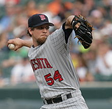 A man wearing a navy blue cap and gray baseball uniform with red lettering throwing a baseball