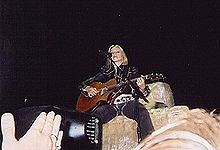 A blond woman sitting on a block of hay and playing a guitar, on a stage. She has short hair and wears grey colored cowboy clothes. Close-up of hands from the audience are visible.