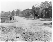 A grayscale view of a rural dirt road with a building visible to the right