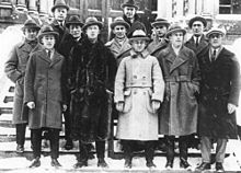 Twelve men pose on the steps in front of a building. They are wearing suits, long jackets and hats.
