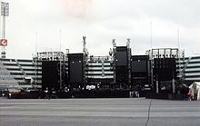 An elaborate concert stage, seen during the day in an empty stadium. The stage comprises several dark, rectangular structures.