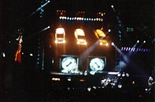 An elaborate concert stage at night. Three cars hang at the stage's rear shining lights towards the performance. Video screens are located behind and to the sides of the stage.
