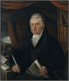 An image of Thomas Coke, Edward Coke's descendant. He is seated at a table with a pen in one hand and a scroll in the other. There are bookshelves in the background.