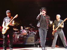 U2 performing on a concert stage. The Edge and Adam Clayton, playing guitars, flank Bono in the foreground, while Larry Mullen, Jr. is behind a drum kit in the background.